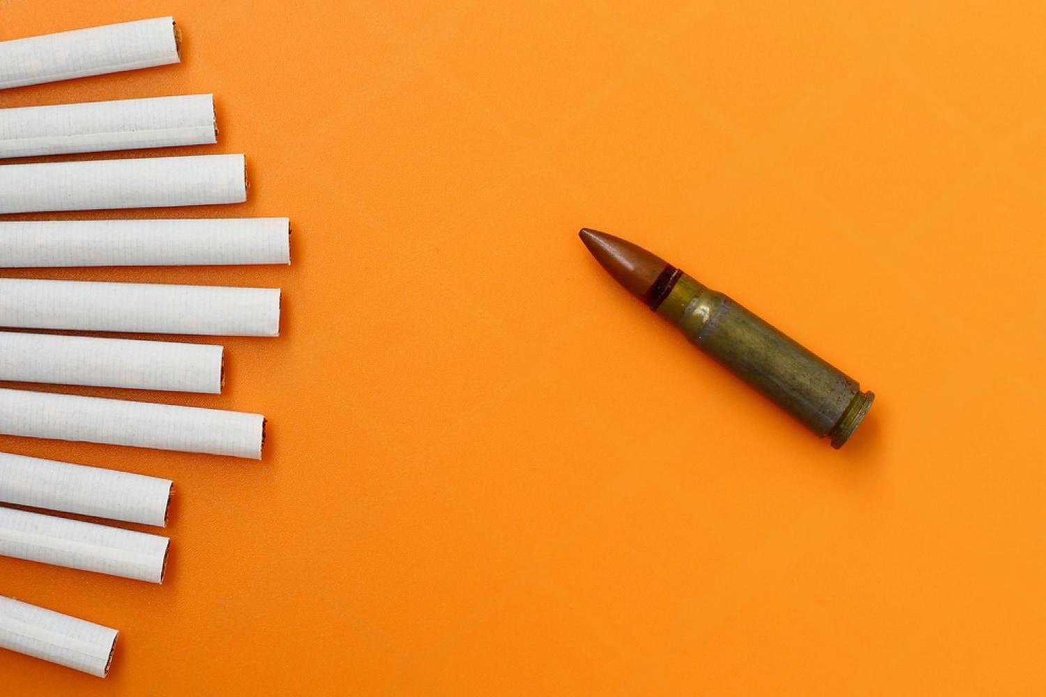 Row of white cigarettes next to a single bullet on an orange background symbolising tobacco risk
